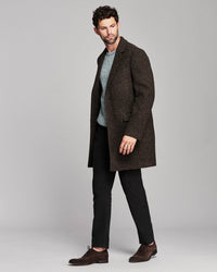 A man modeling a dark brown and black houndstooth patterned overcoat, worn open over a gray knit sweater, black trousers, and brown suede shoes, against a solid light gray studio background.