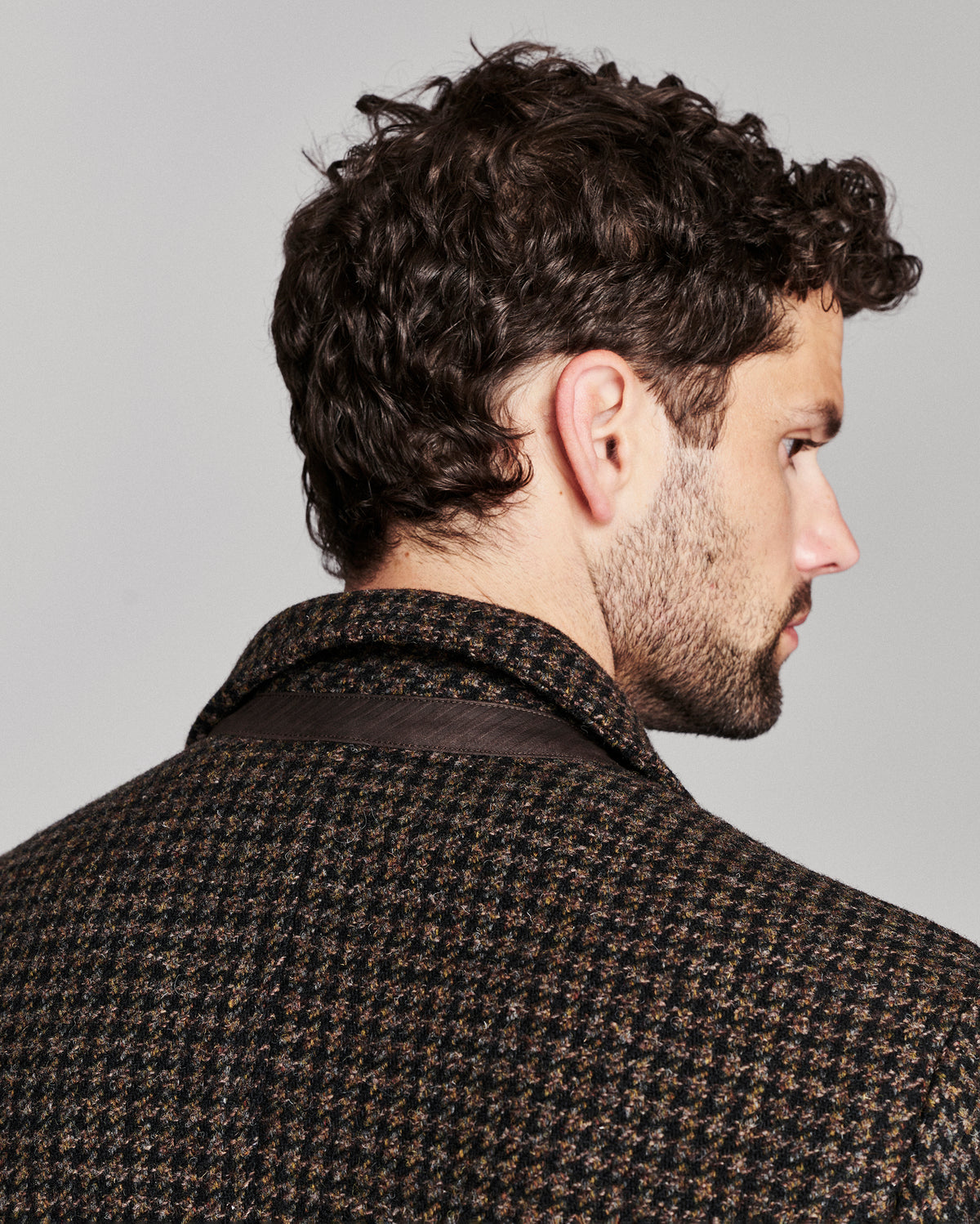 A man shown from the back and side profile wearing a dark brown and black houndstooth patterned jacket against a light gray studio background.