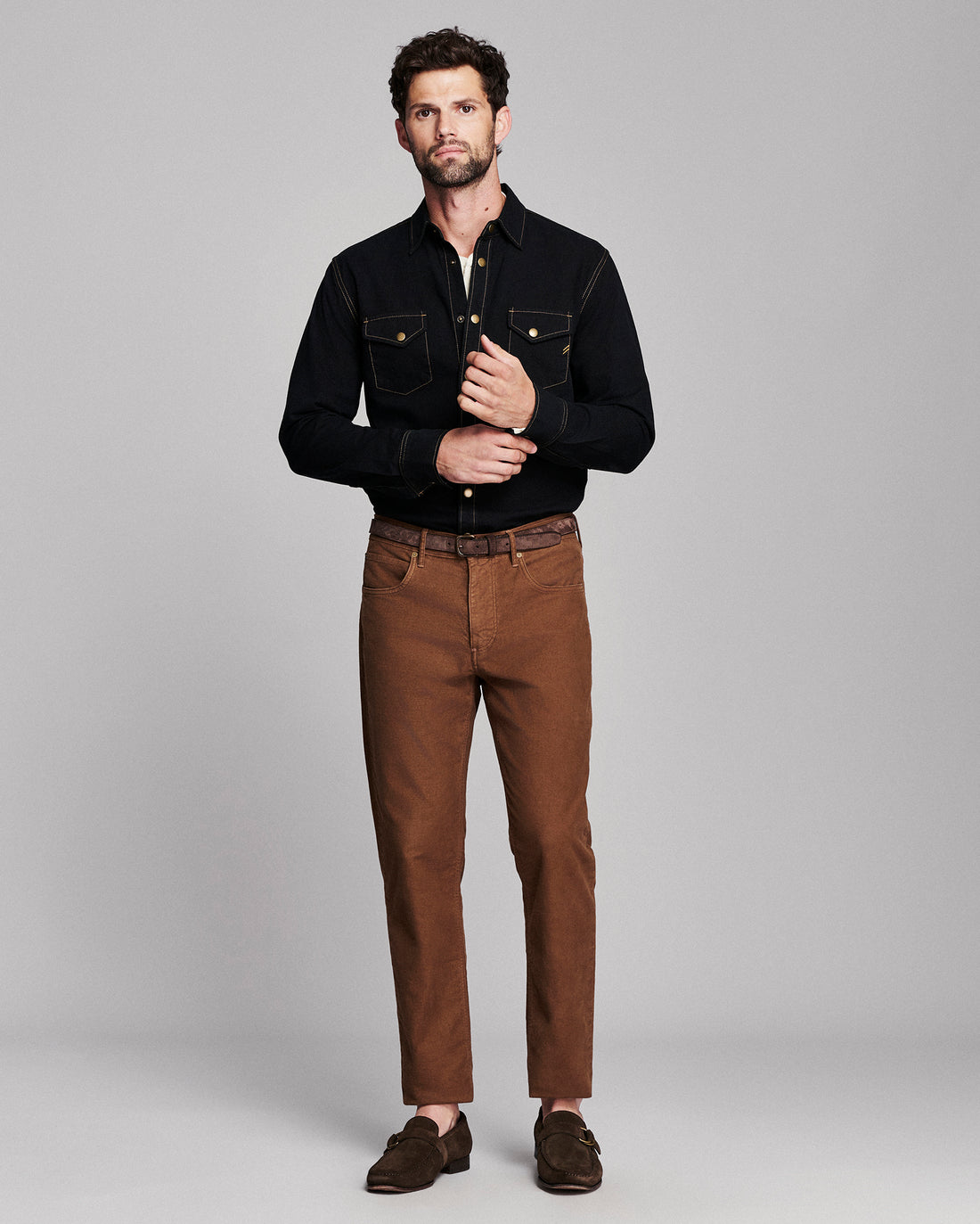 A man modeling a black long-sleeve button-up shirt with chest pockets and brown trousers, standing against a light gray studio background.