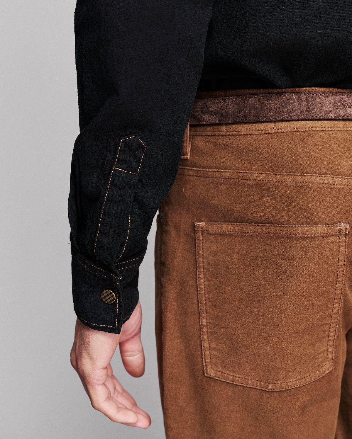 Close-up of a person wearing brown corduroy trousers with a back pocket, a black shirt, and a brown belt, with the hand and sleeve of the shirt visible against a gray background.
