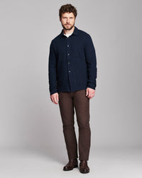 A man modeling a dark navy blue long-sleeve button-up shirt, paired with brown trousers and dark brown boots, against a solid gray studio background.