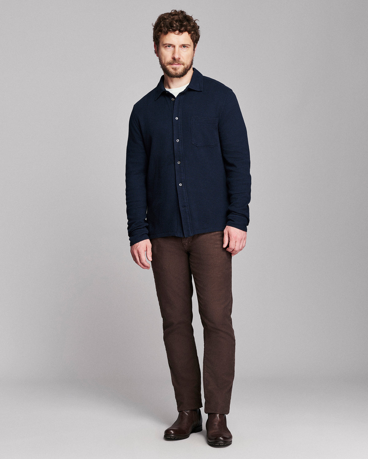 A man modeling a dark navy blue long-sleeve button-up shirt, paired with brown trousers and dark brown boots, against a solid gray studio background.