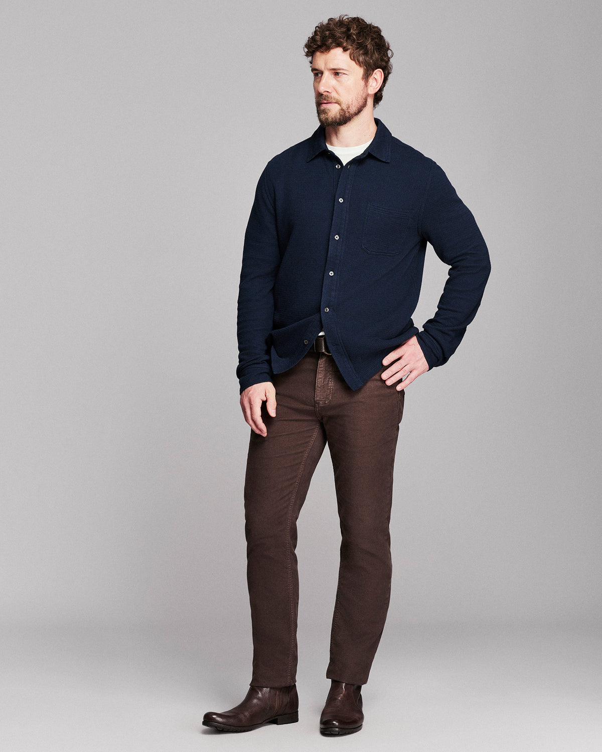 A man modeling a dark navy blue button-down shirt over a white undershirt, paired with brown trousers and dark brown leather boots, against a solid gray studio background.