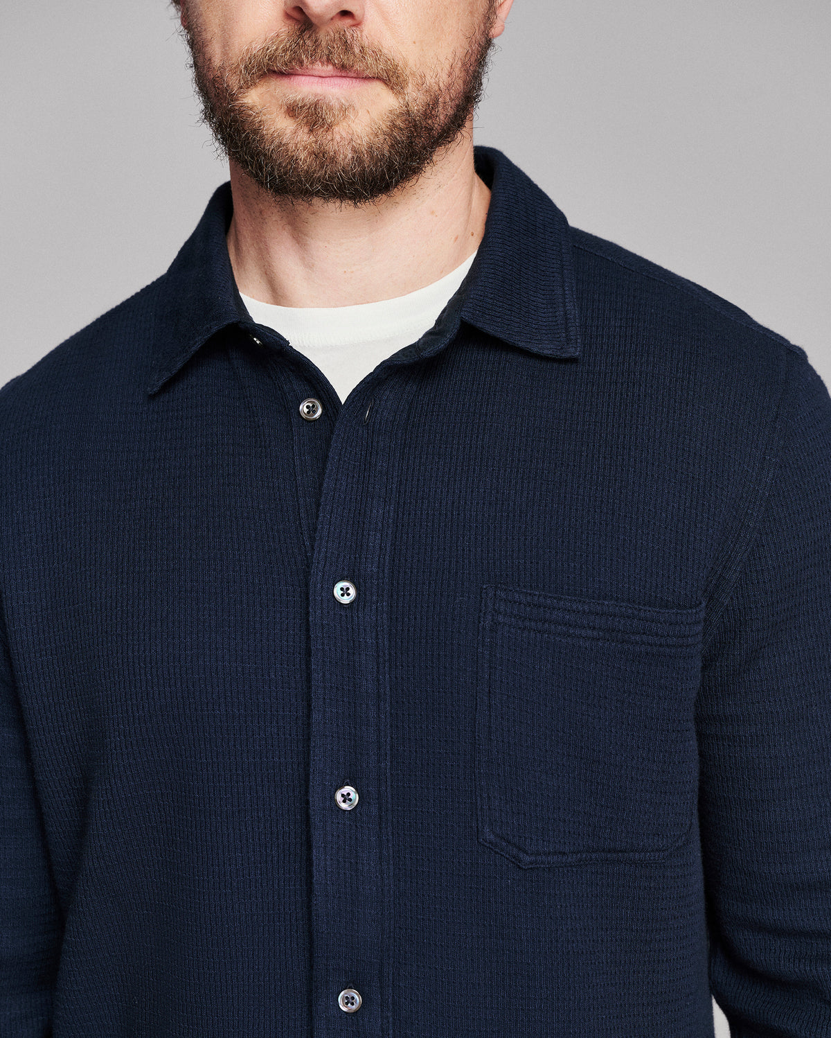 A close-up of a man wearing a dark navy blue textured button-up shirt over a white crew-neck undershirt, against a solid gray background.