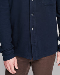 A close-up of a person wearing a dark navy blue waffle-knit button-up shirt with brown pants, showing the chest, buttons, and hands.