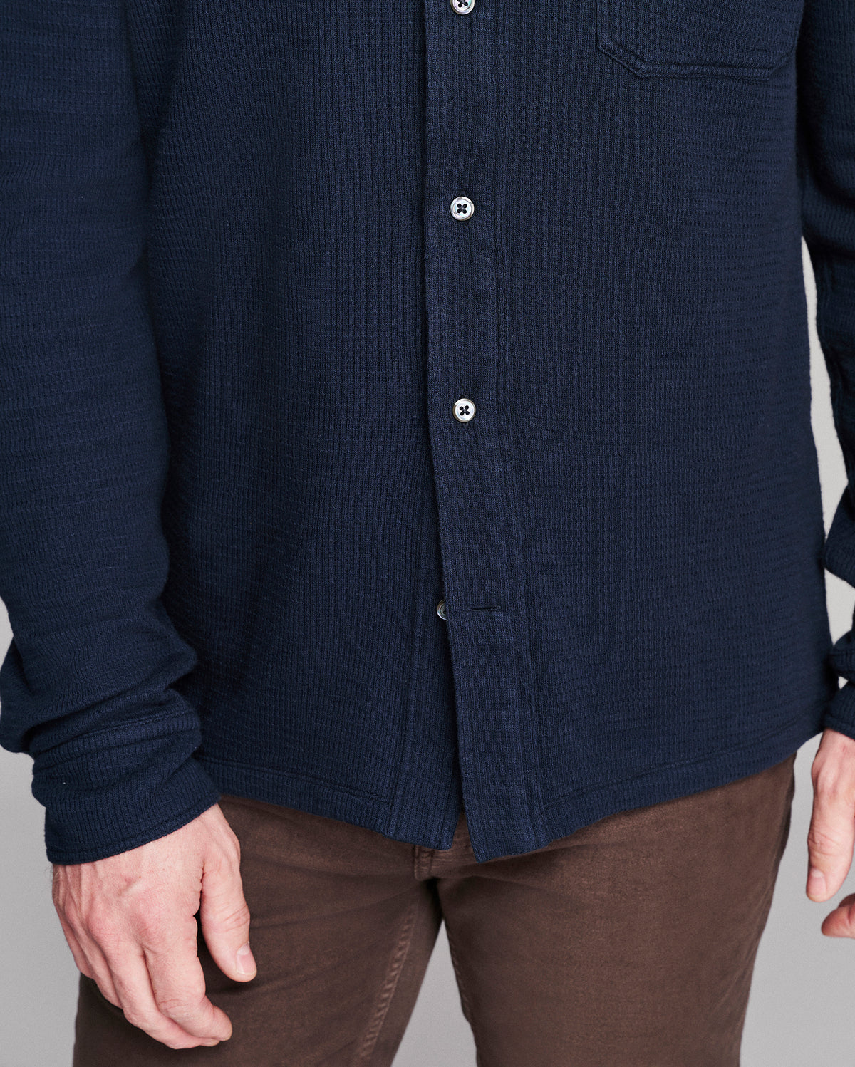 A close-up of a person wearing a dark navy blue waffle-knit button-up shirt with brown pants, showing the chest, buttons, and hands.
