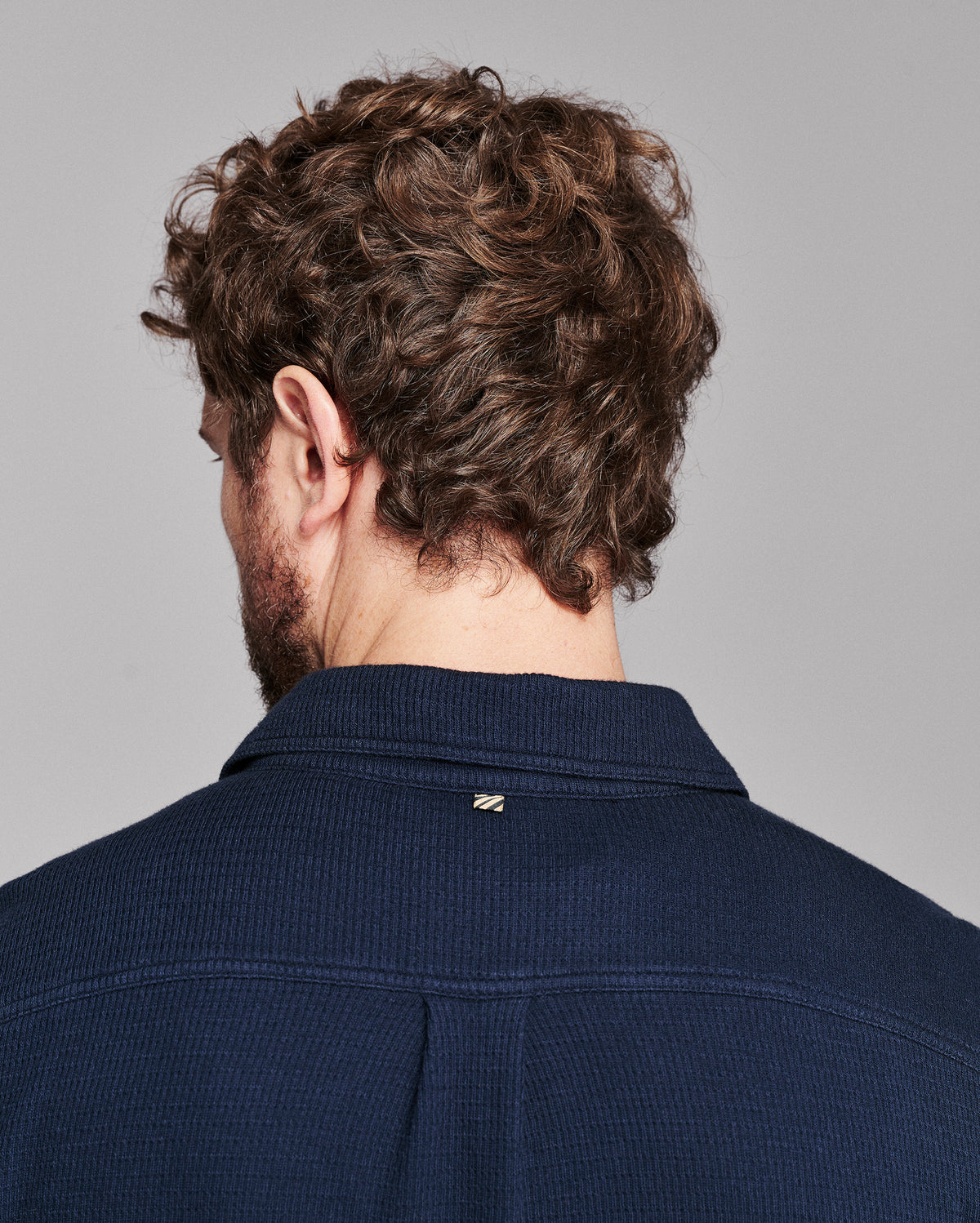 Back view of a man with curly brown hair wearing a dark navy ribbed knit collared shirt against a solid gray background.