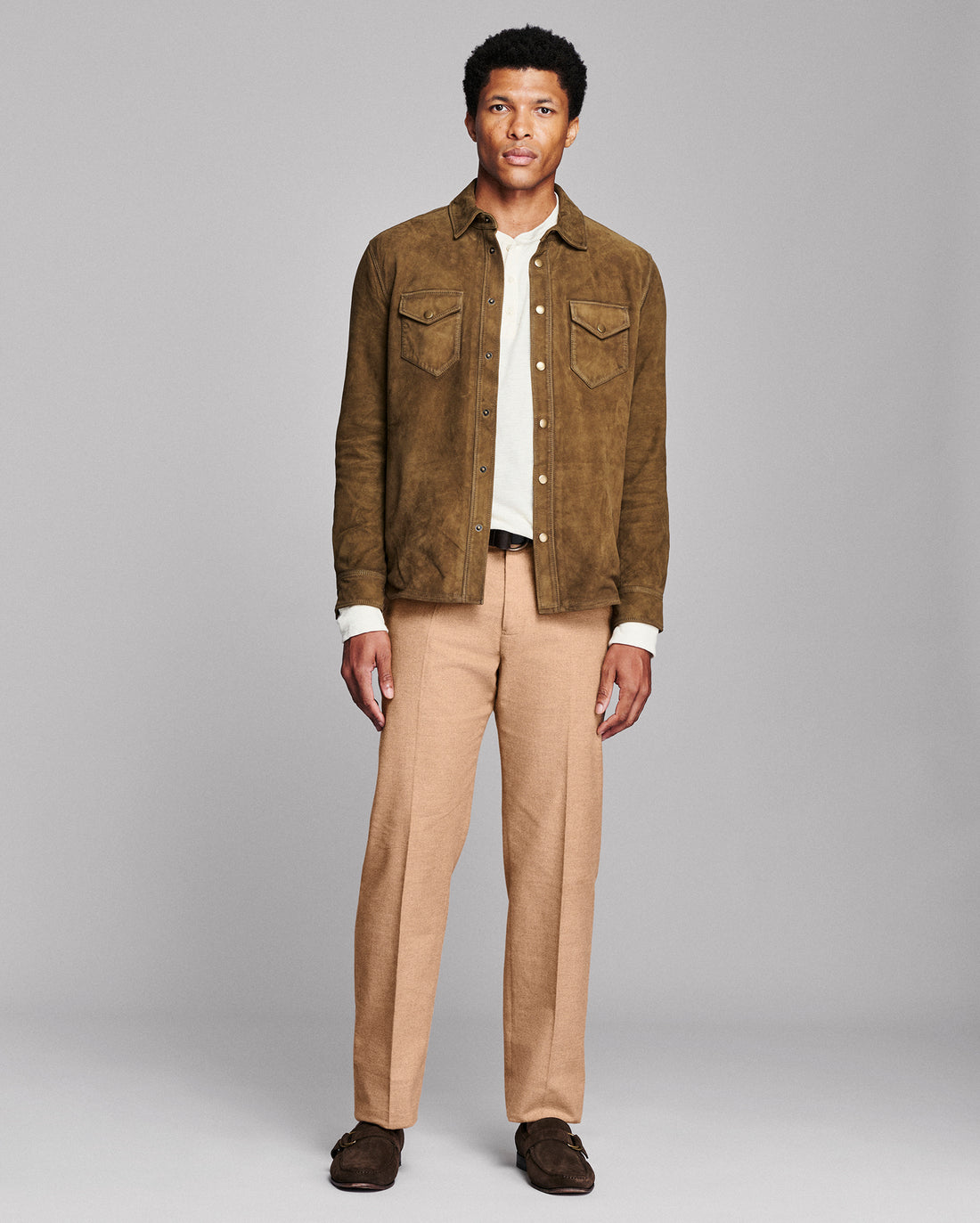 A man modeling a brown suede jacket over a white shirt, paired with tan trousers and dark brown loafers, against a solid gray studio background.