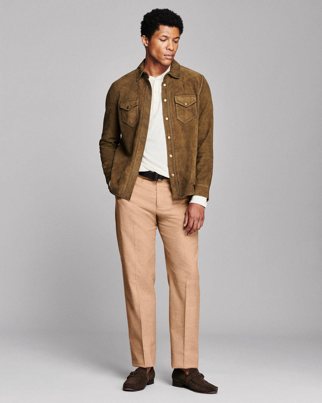 A man modeling a brown suede button-up shirt jacket over a white t-shirt, paired with beige trousers and dark brown suede loafers, against a solid gray studio background.