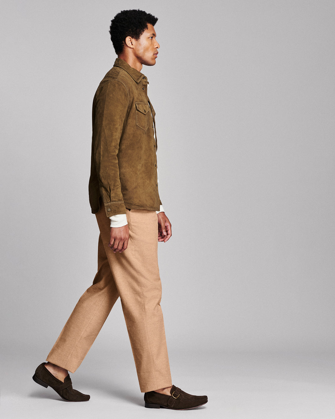 A man in side profile wearing a brown suede jacket, beige trousers, and dark brown loafers, walking against a solid light gray studio background.