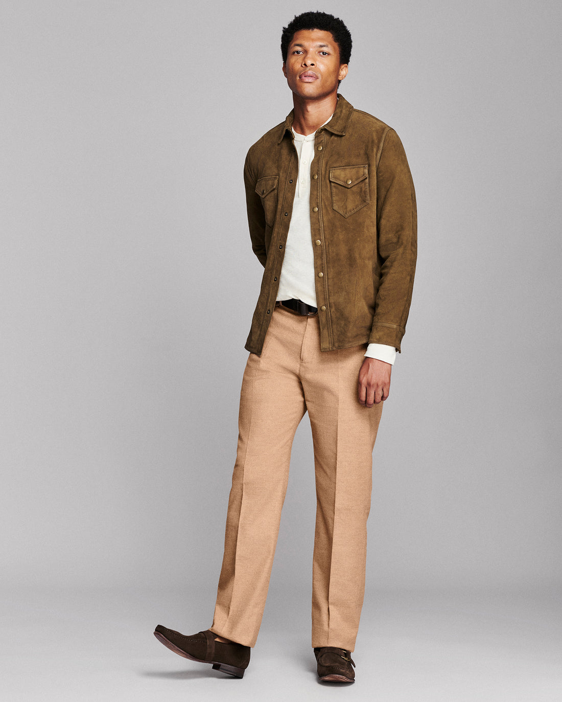 A man modeling a brown suede jacket over a white shirt, paired with tan trousers and dark brown loafers, against a solid gray studio background.