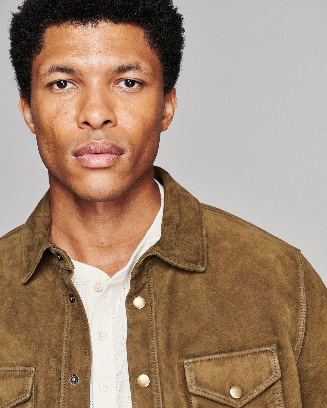 A close-up portrait of a man wearing a brown suede button-up shirt jacket over a white henley shirt, set against a solid gray studio background.