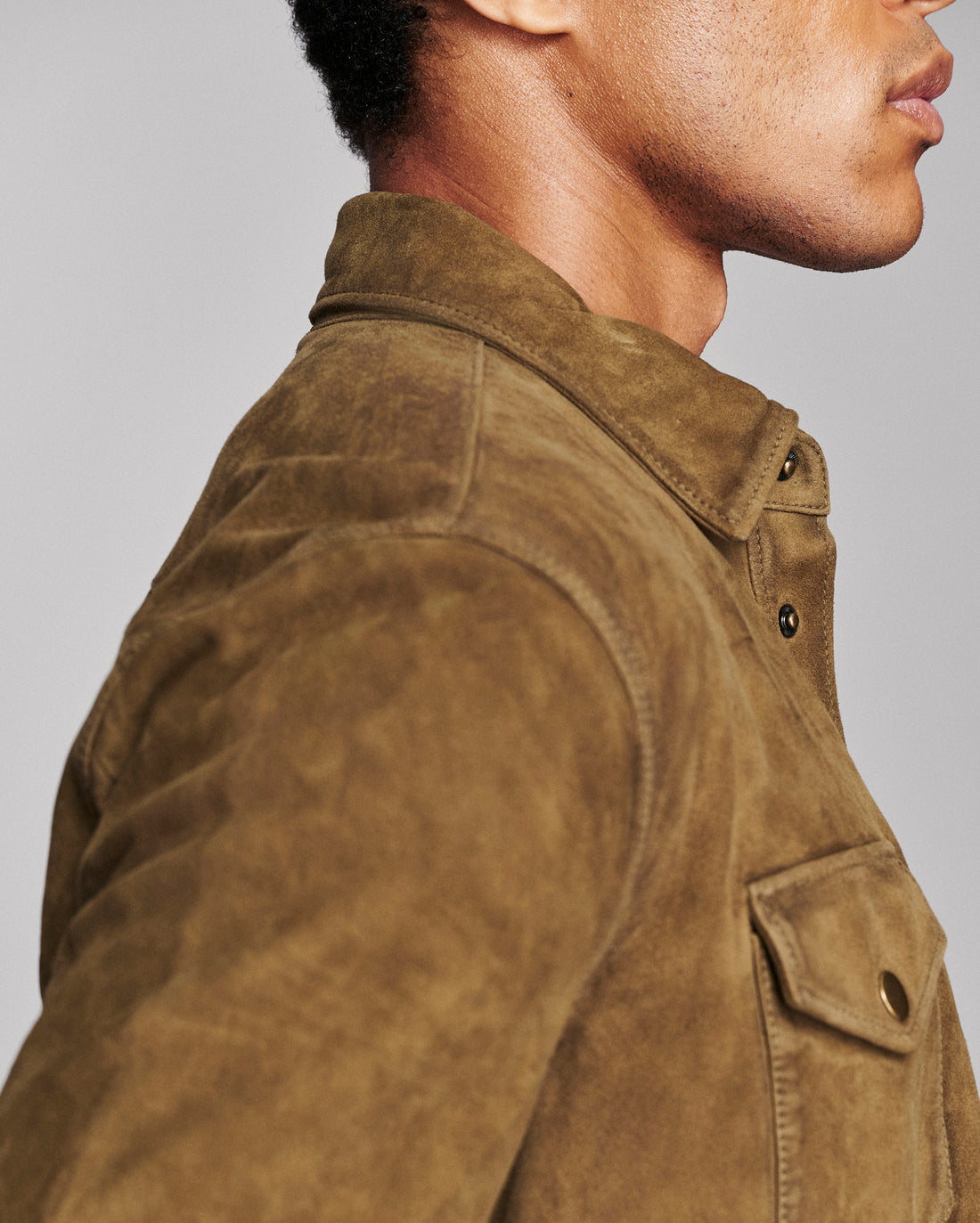 Close-up side profile of a person wearing a brown suede or nubuck overshirt with a collar and flap chest pocket, against a light gray studio background.