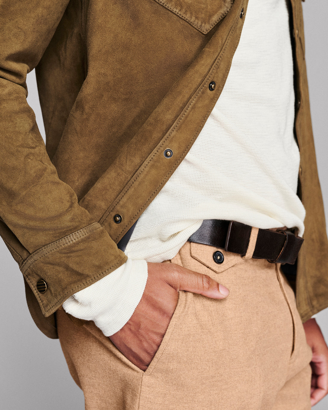A close-up of a person wearing a brown suede jacket over a white shirt, paired with tan trousers and a dark belt, against a plain light gray background.