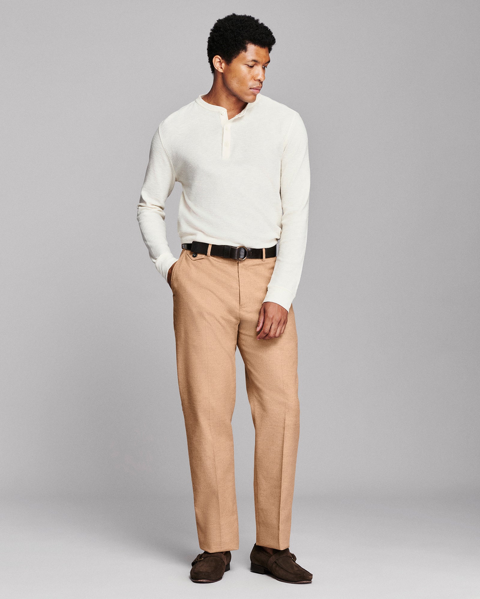 A man modeling a cream long-sleeve henley shirt and tan trousers with a black belt, standing against a solid gray studio background.