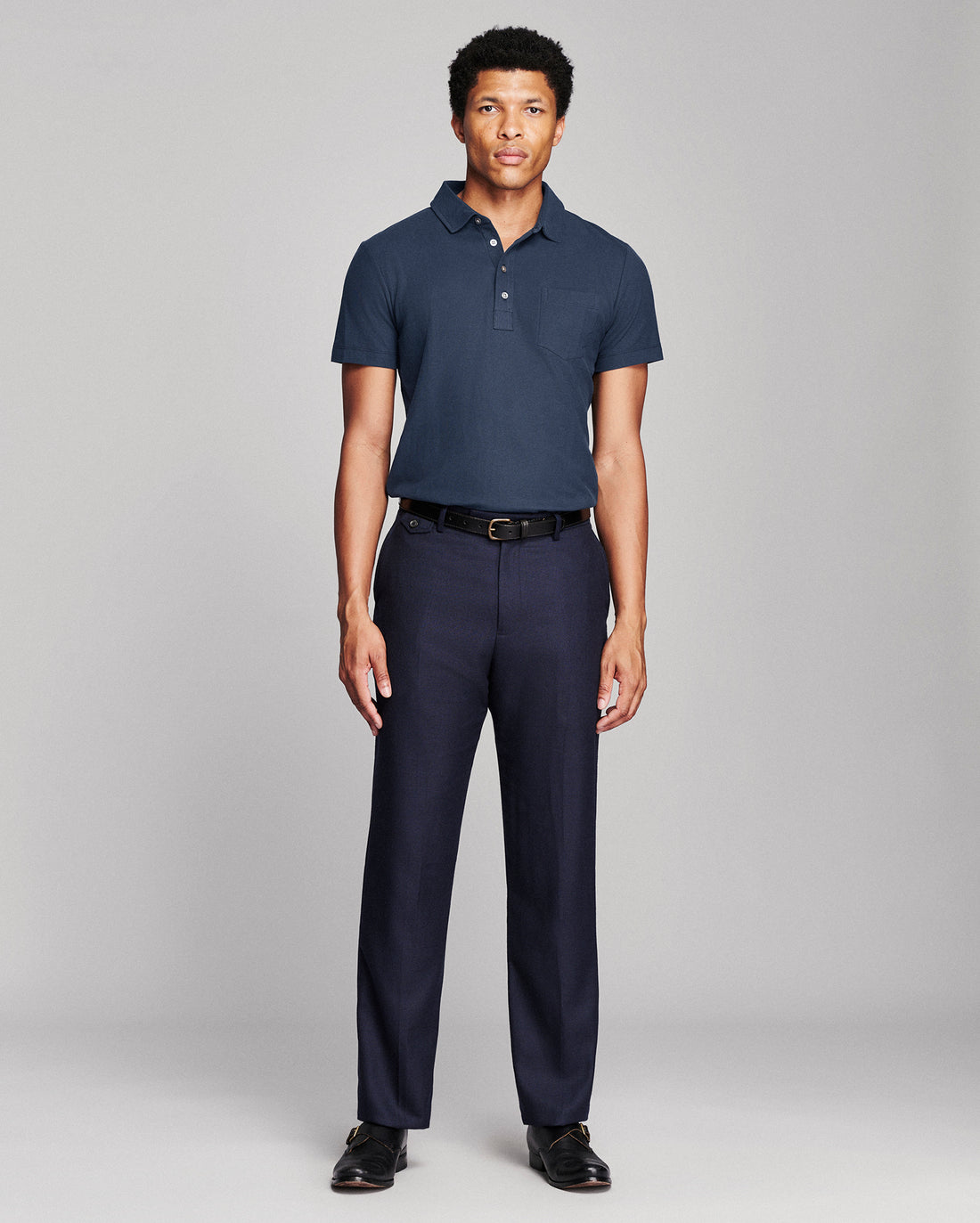A man modeling a navy blue short-sleeve polo shirt and matching navy trousers with a black belt, standing against a light gray studio background.