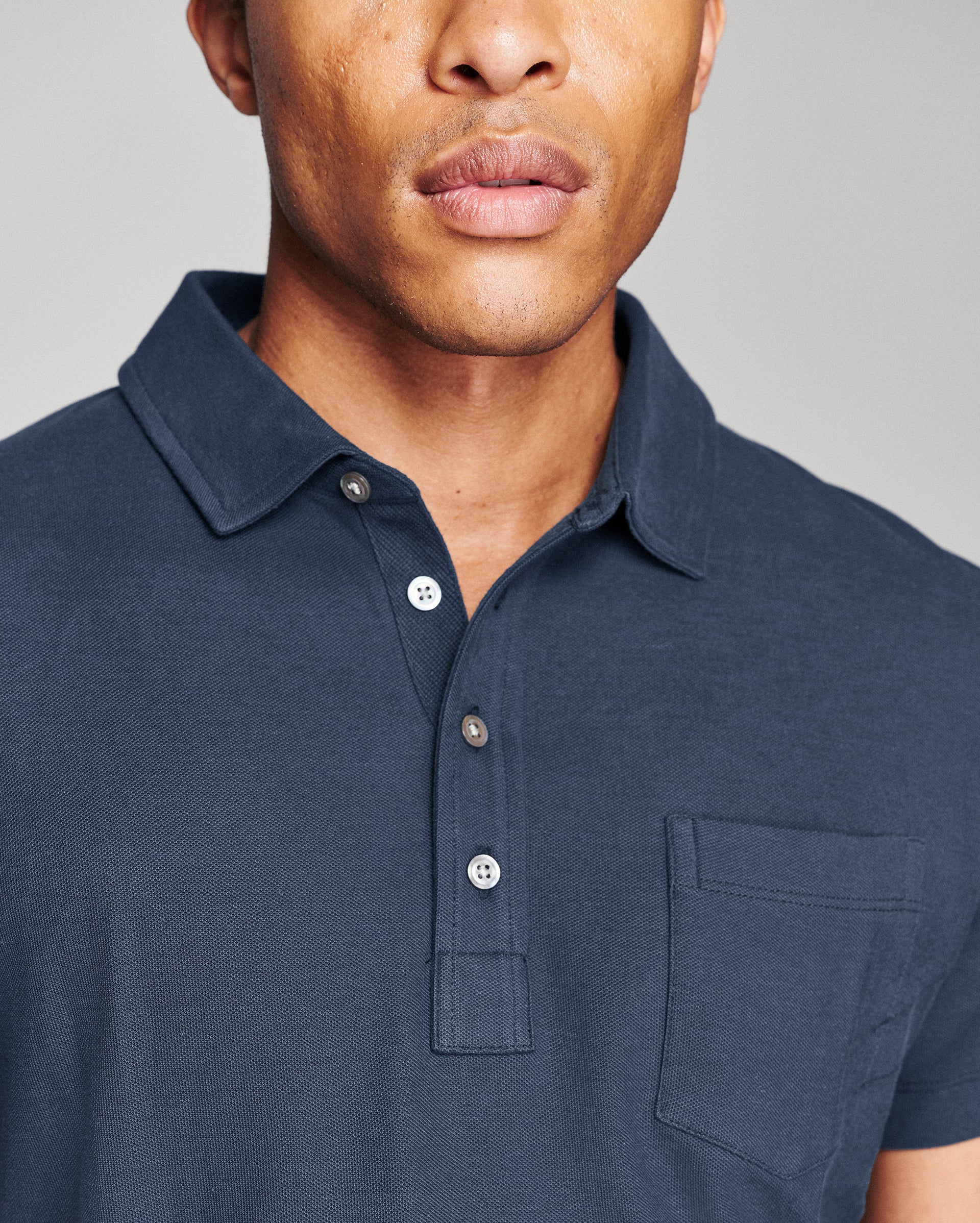 Close-up of a man wearing a dark navy blue short-sleeve polo shirt with a collar, three-button placket, and a chest pocket, set against a plain light gray background.