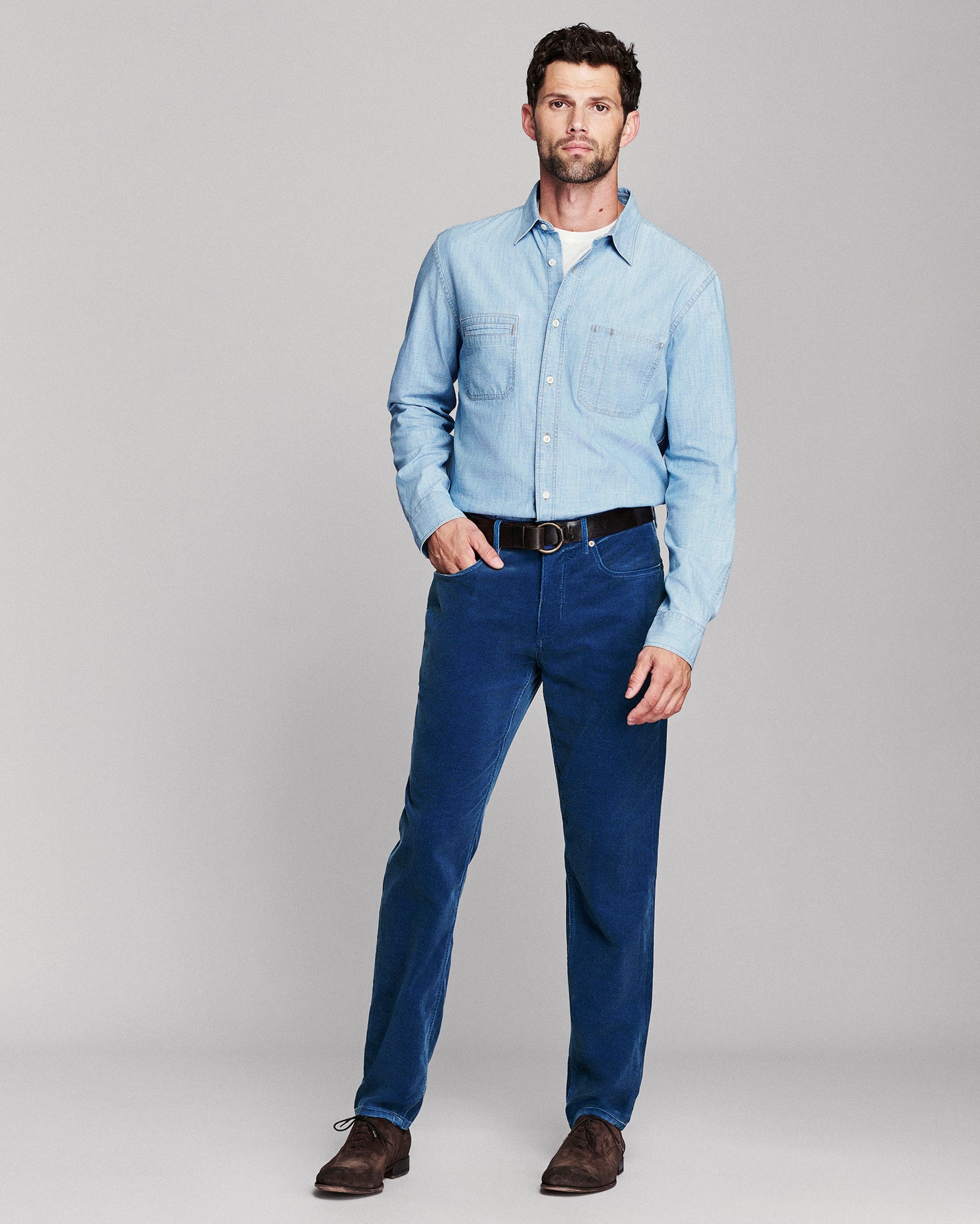 A man modeling a light blue long-sleeve button-up shirt and dark blue jeans with a black belt, standing against a neutral gray studio background.