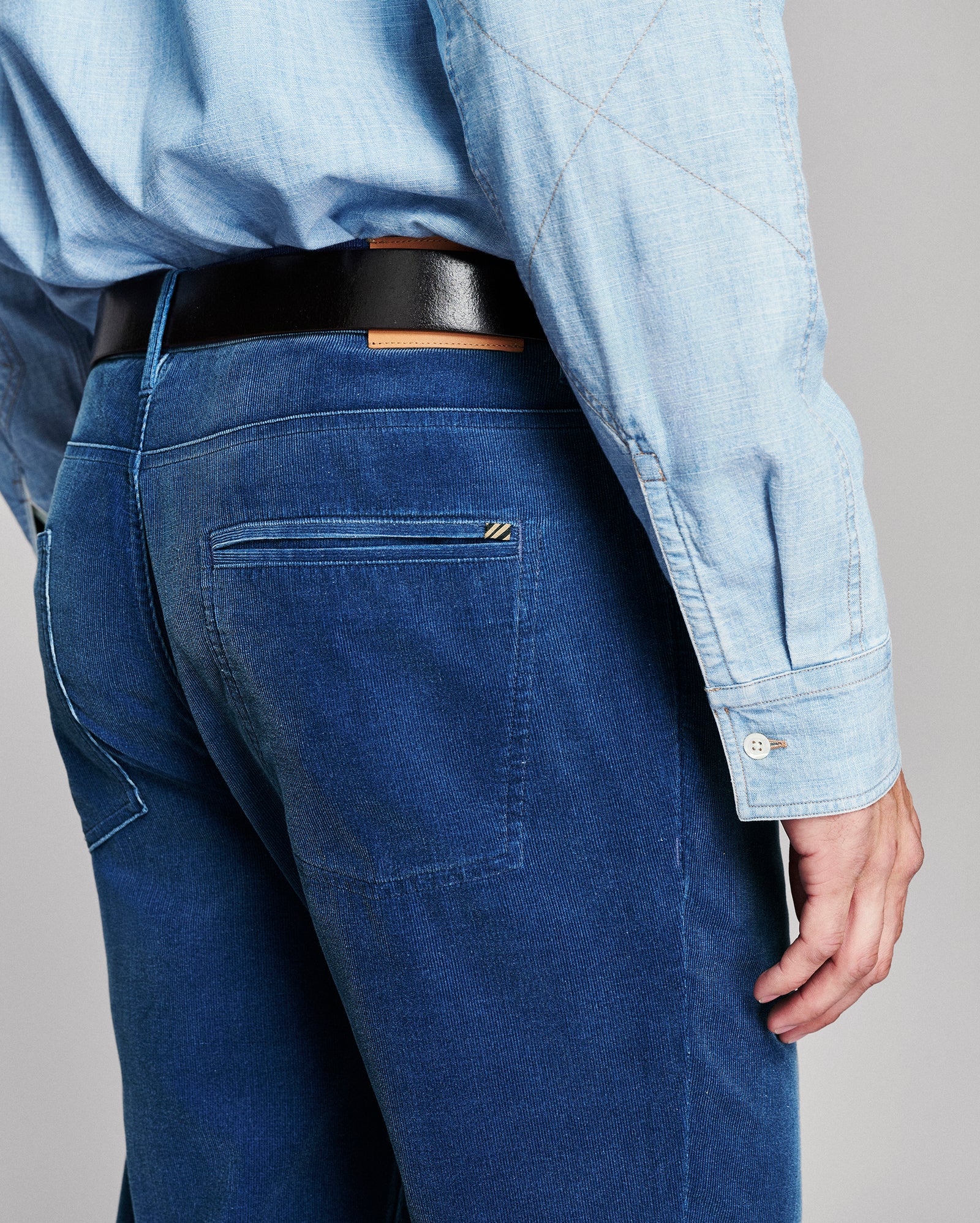 Close-up back view of a person wearing dark blue jeans, a light blue shirt, and a black belt against a plain light gray background.