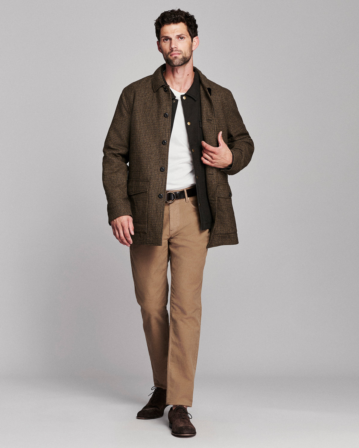 A man modeling a brown textured jacket over a white shirt and black vest, paired with tan trousers and dark brown shoes, against a solid gray studio background.