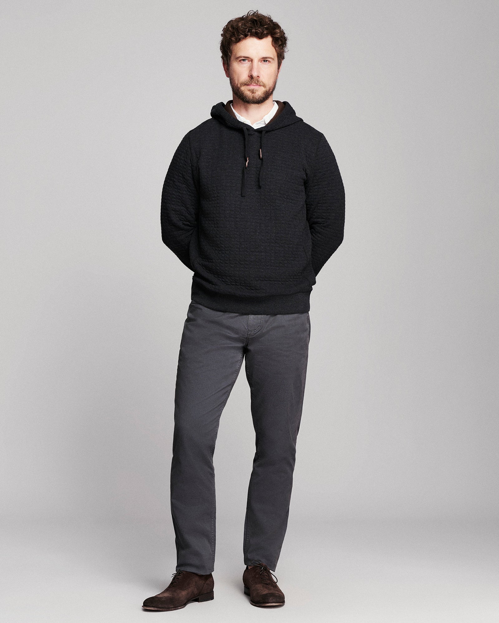 A man modeling a black textured knit hoodie with gray trousers and brown shoes, standing against a light gray studio background.