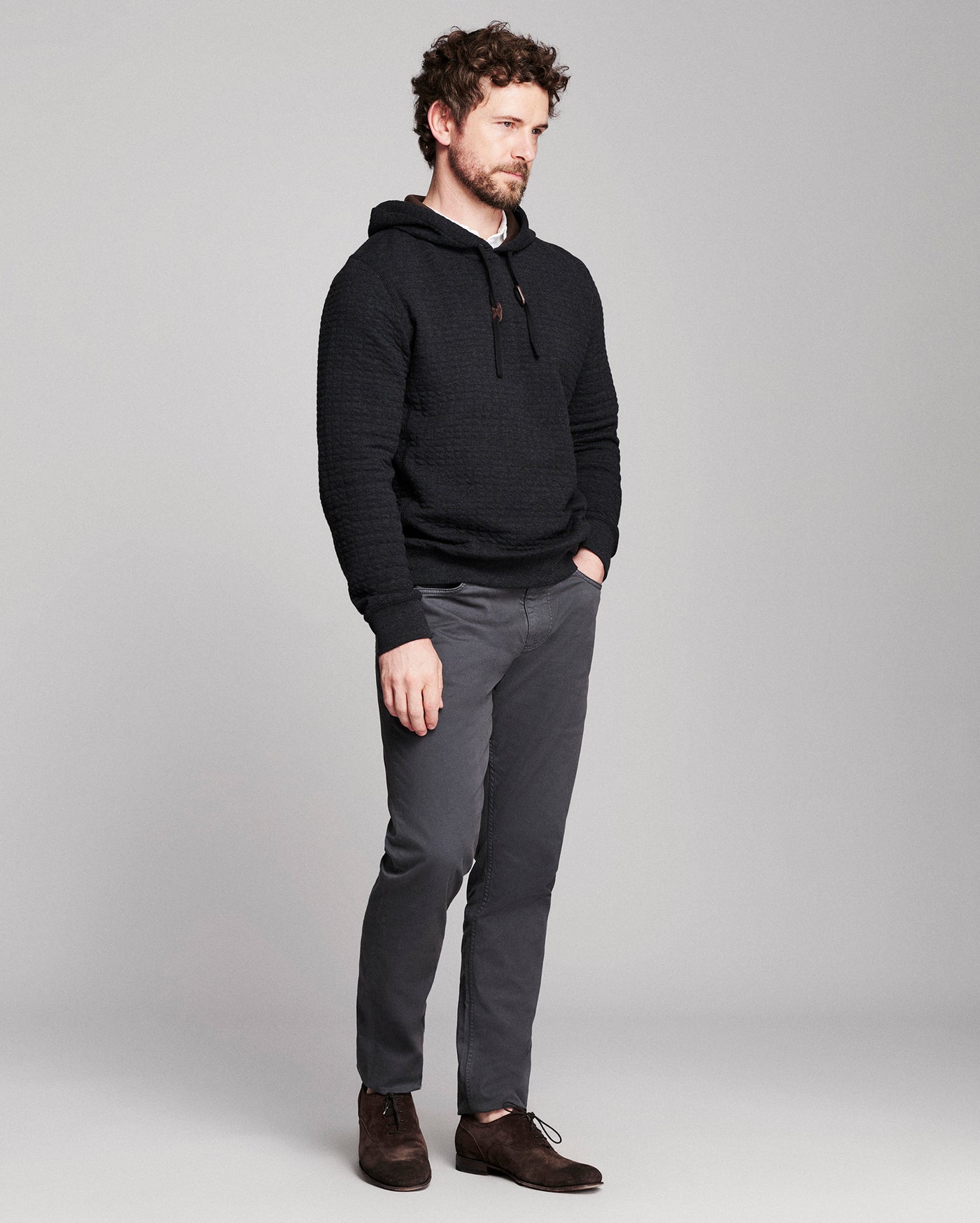 A man wearing a black textured pullover hoodie, dark gray slim-fit trousers, and brown leather shoes, standing against a light gray studio background.