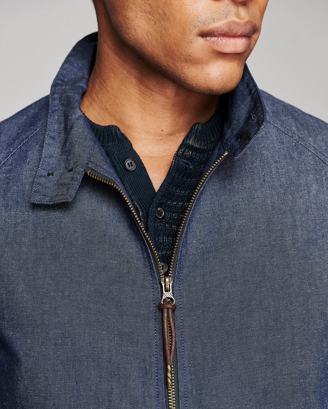 Close-up of a man wearing a blue textured zip-up jacket over a dark navy knit shirt, showing the collar, zipper, and fabric details against a neutral gray background.