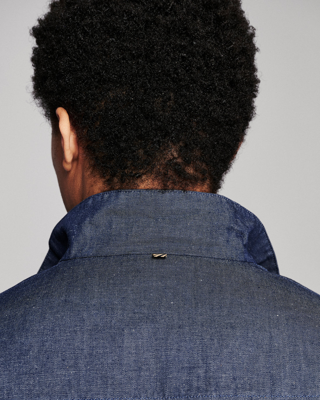 Back view of a person with short dark curly hair wearing a textured blue denim or chambray collared shirt jacket against a plain light gray background.