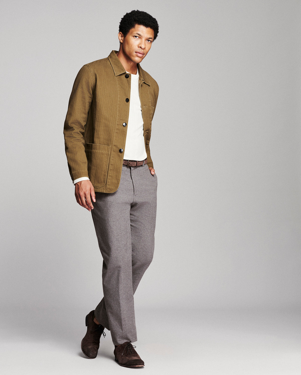 A man modeling a brown corduroy jacket over a white t-shirt, paired with gray trousers and brown suede shoes, against a solid gray studio background.