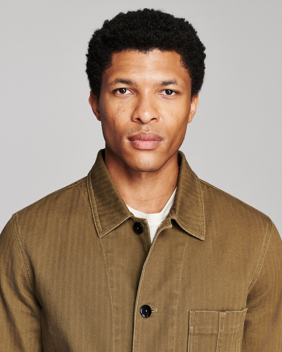 A man wearing a brown corduroy button-up shirt over a white undershirt, photographed against a solid gray background.