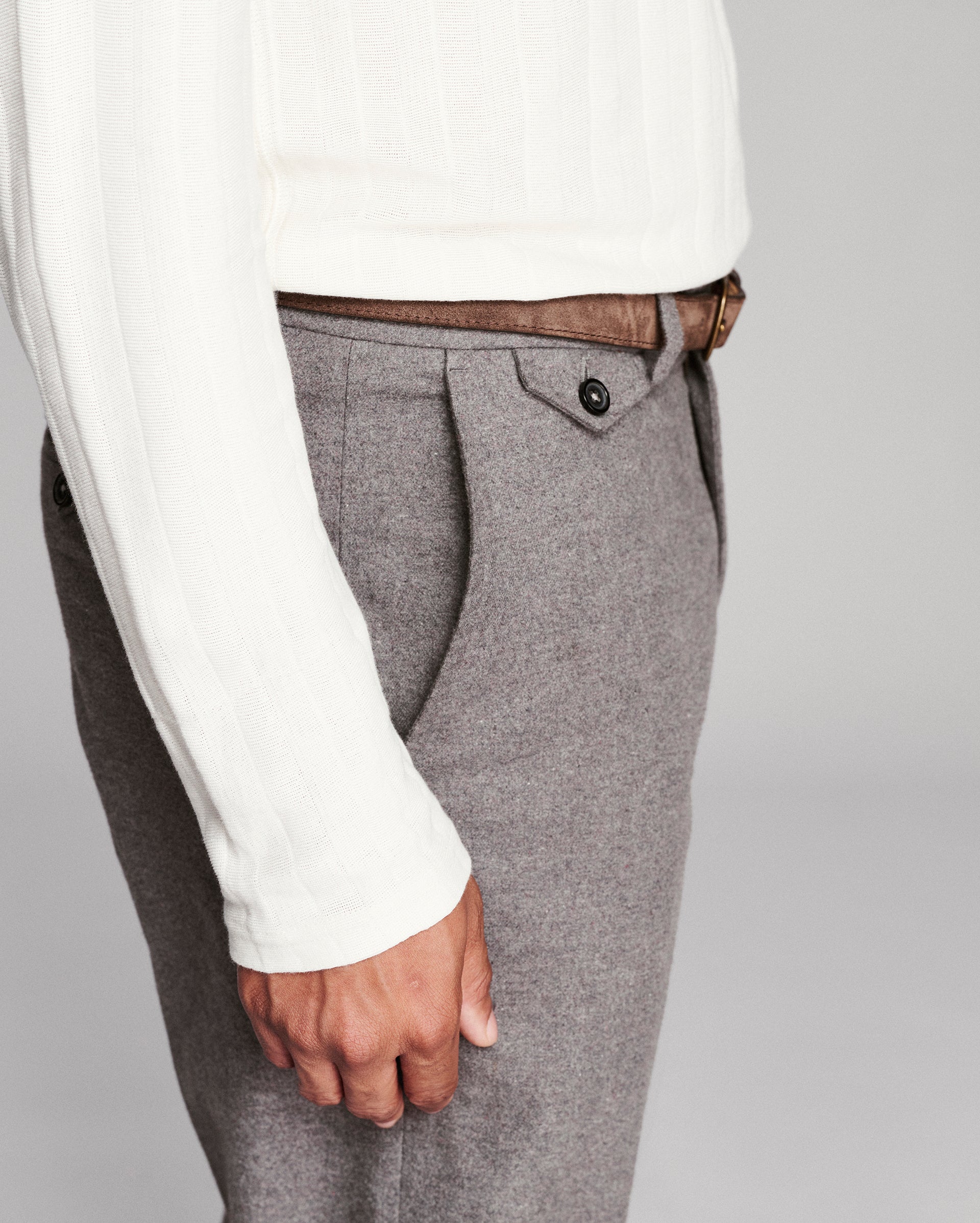 Close-up of a person wearing gray textured trousers with a brown belt and a white ribbed sweater, shown against a light gray studio background.