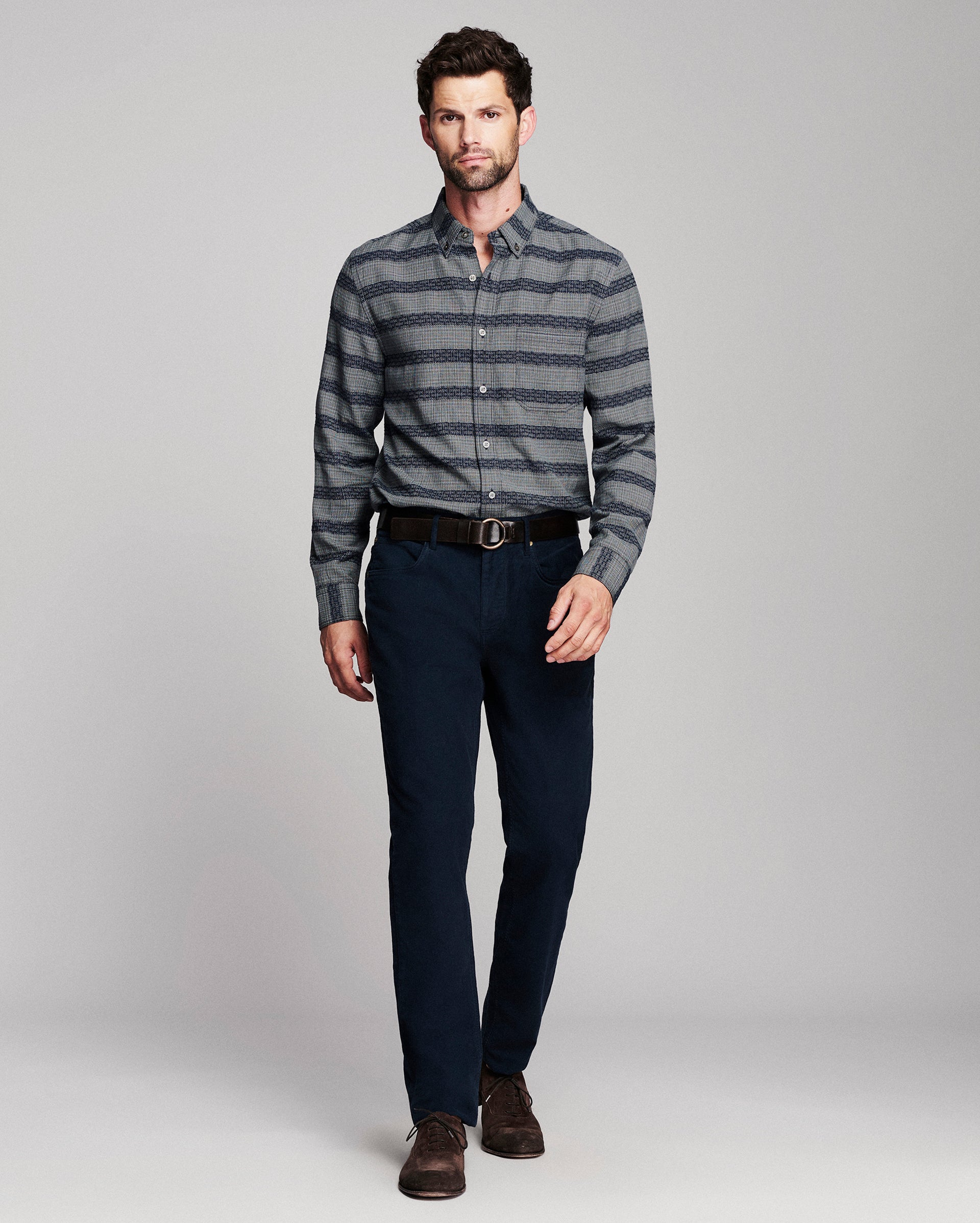A man modeling a gray and black striped long-sleeve button-down shirt, paired with dark navy trousers, a black belt, and brown dress shoes, against a light gray studio background.