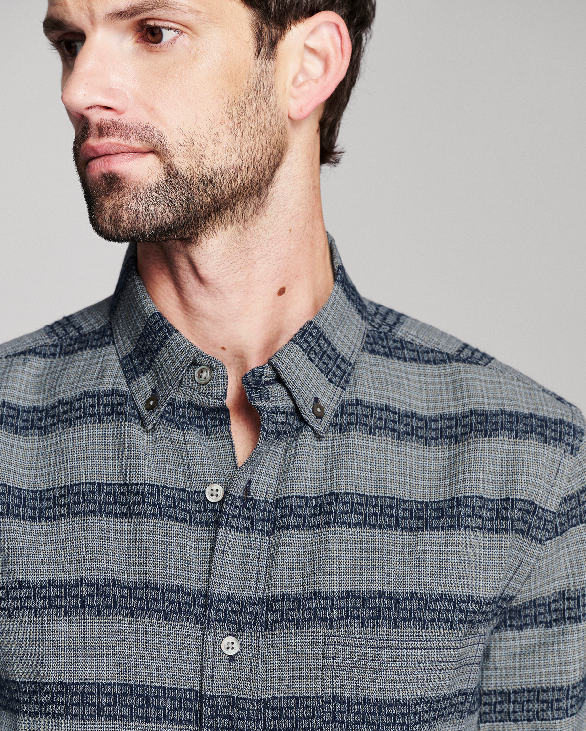 A close-up of a man wearing a blue and gray striped button-down shirt with a subtle geometric pattern.