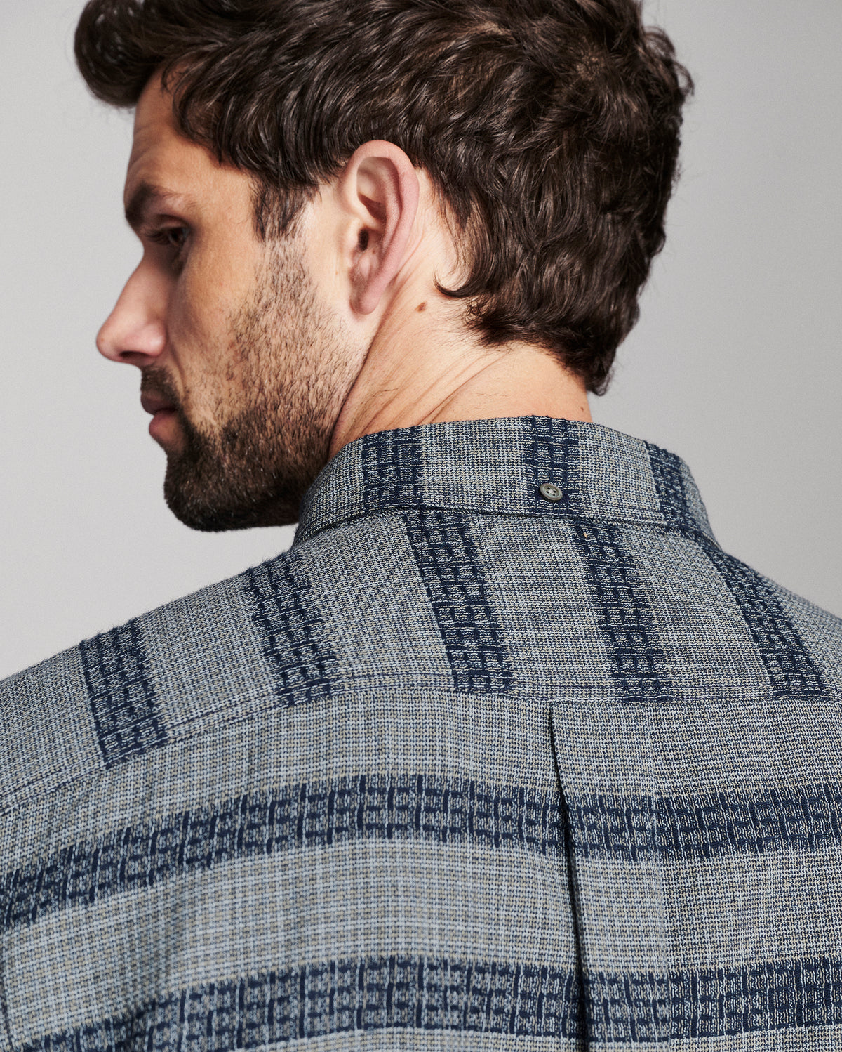 Close-up side profile of a man wearing a textured blue and gray patterned button-down shirt, showing the collar and fabric weave against a neutral gray background.