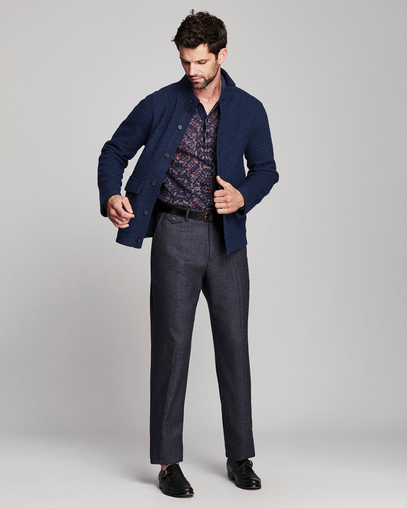 A man modeling a navy blue knit cardigan over a patterned shirt, paired with dark charcoal trousers and black loafers, against a light gray studio background.