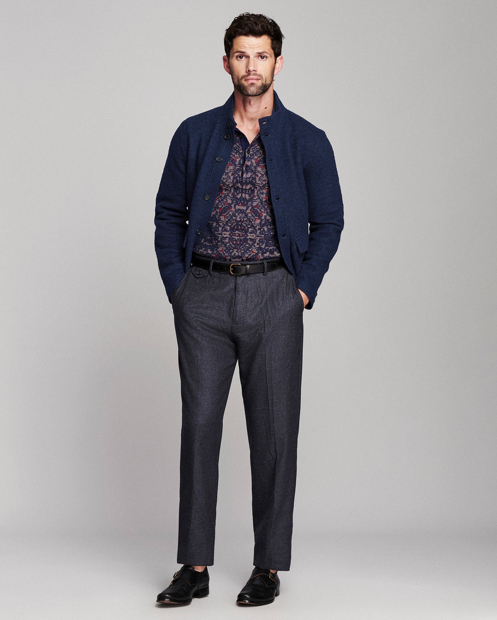 A man modeling a dark navy blue knit cardigan over a patterned button-down shirt, paired with dark gray trousers and black dress shoes, against a light gray studio background.