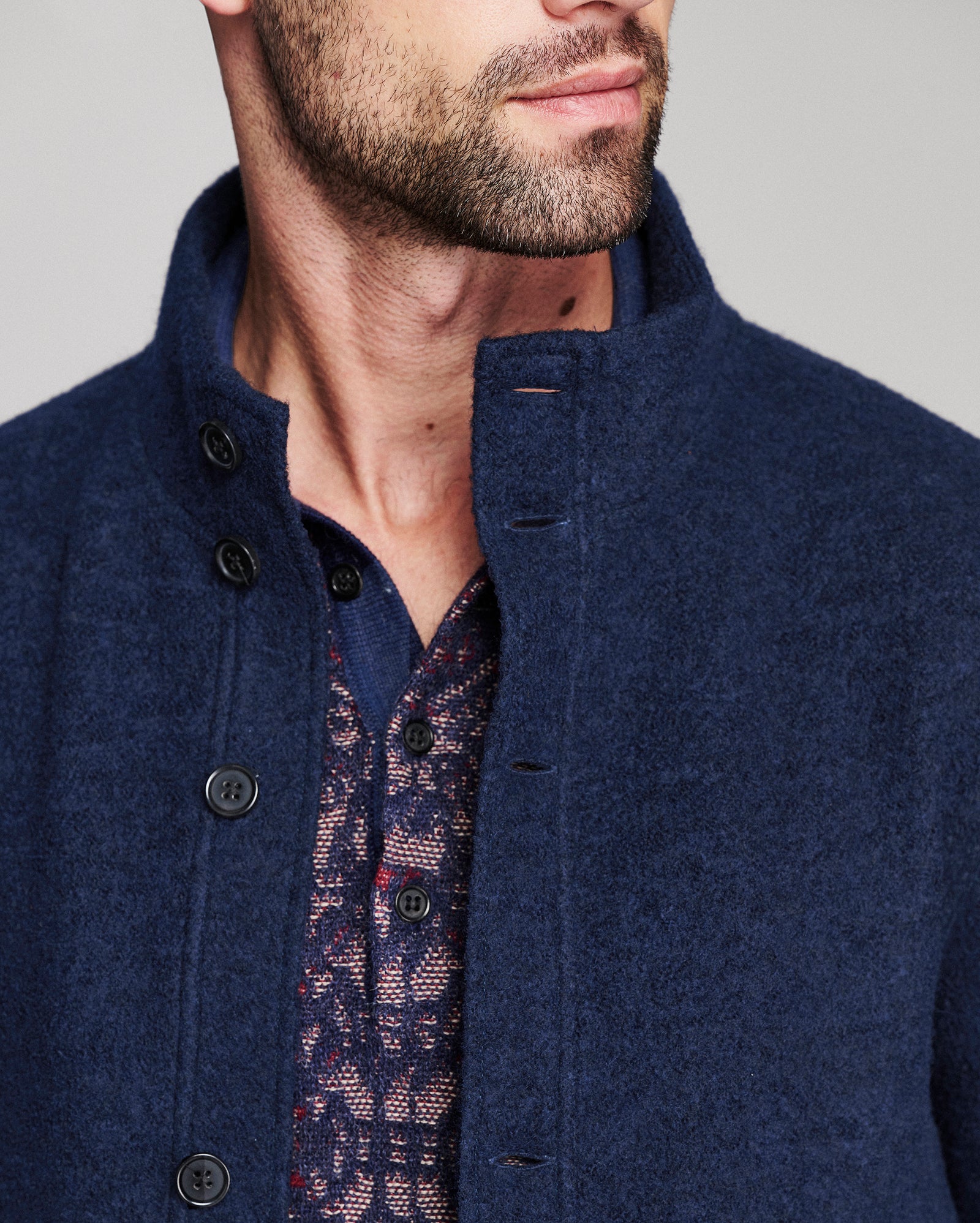 Close-up of a man wearing a navy blue textured wool-blend jacket with a high stand collar and dark buttons, layered over a patterned shirt.