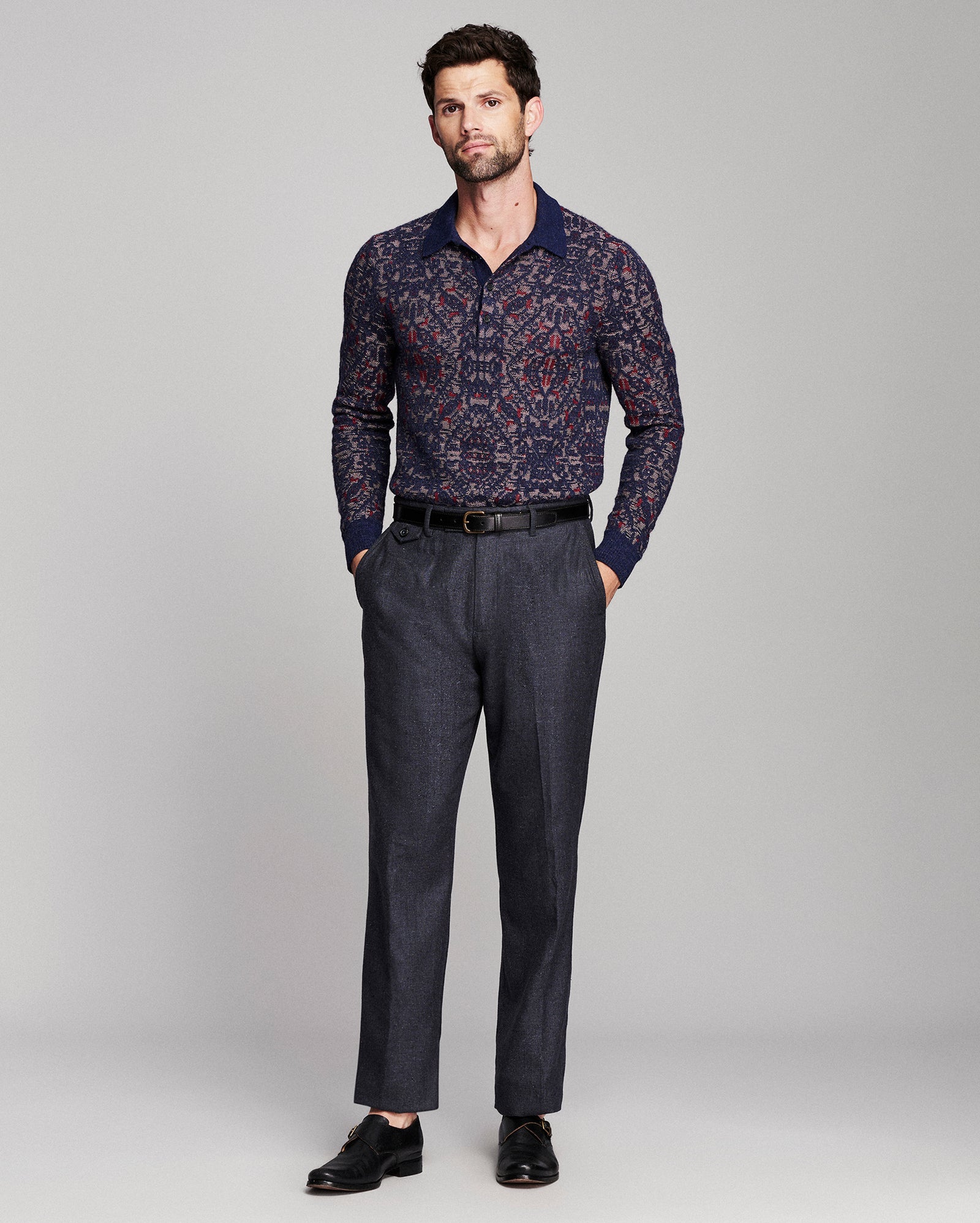 A man modeling a long-sleeve button-up shirt with a navy blue and red geometric pattern, paired with dark gray trousers and black dress shoes, against a light gray studio background.