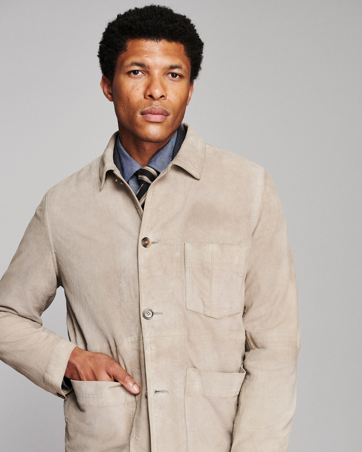 A man wearing a light beige corduroy utility jacket over a blue collared shirt and striped tie, photographed against a solid gray studio background.