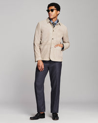 A man modeling a light beige corduroy jacket over a blue patterned shirt, paired with navy trousers and black dress shoes, against a light gray studio background.