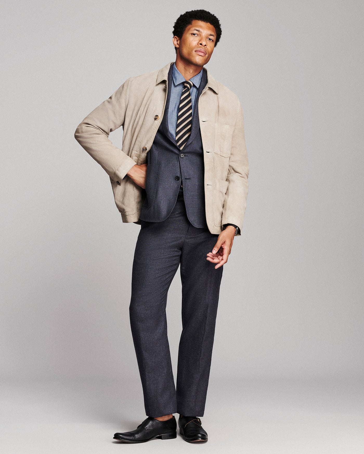 A man modeling a beige overshirt over a navy blue suit, including a blue dress shirt, striped tie, and navy trousers, paired with black leather dress shoes, against a light gray studio background.