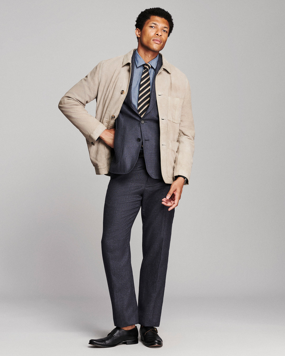 A man modeling a beige overshirt over a navy blue suit, including a blue dress shirt, striped tie, and navy trousers, paired with black leather dress shoes, against a light gray studio background.