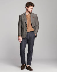 A man modeling a brown textured blazer over a tan knit polo shirt, paired with navy trousers and brown suede loafers, against a solid light gray studio background.