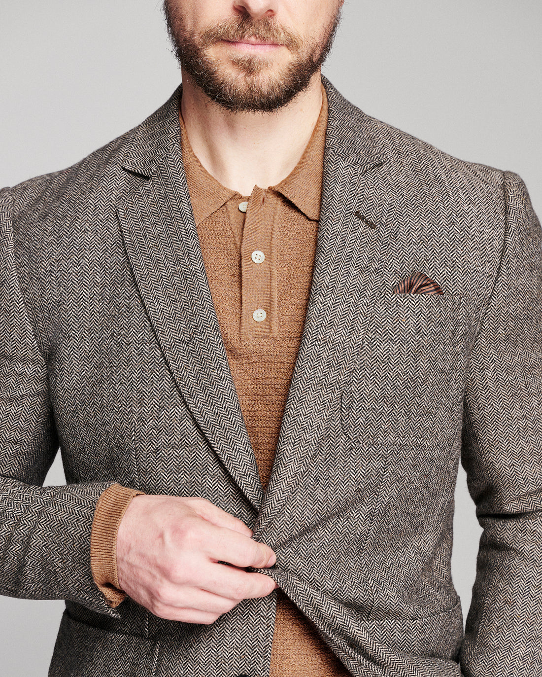 A man wearing a brown and gray herringbone blazer over a brown knit polo shirt, with a matching pocket square, against a solid gray background.