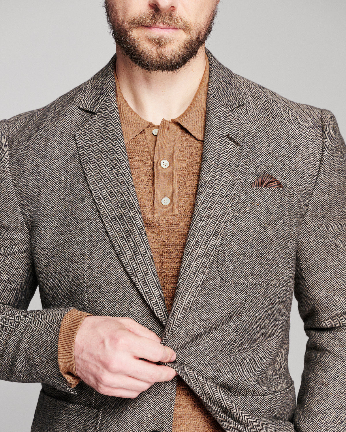 A man wearing a brown and gray herringbone blazer over a brown knit polo shirt, with a matching pocket square, against a solid gray background.