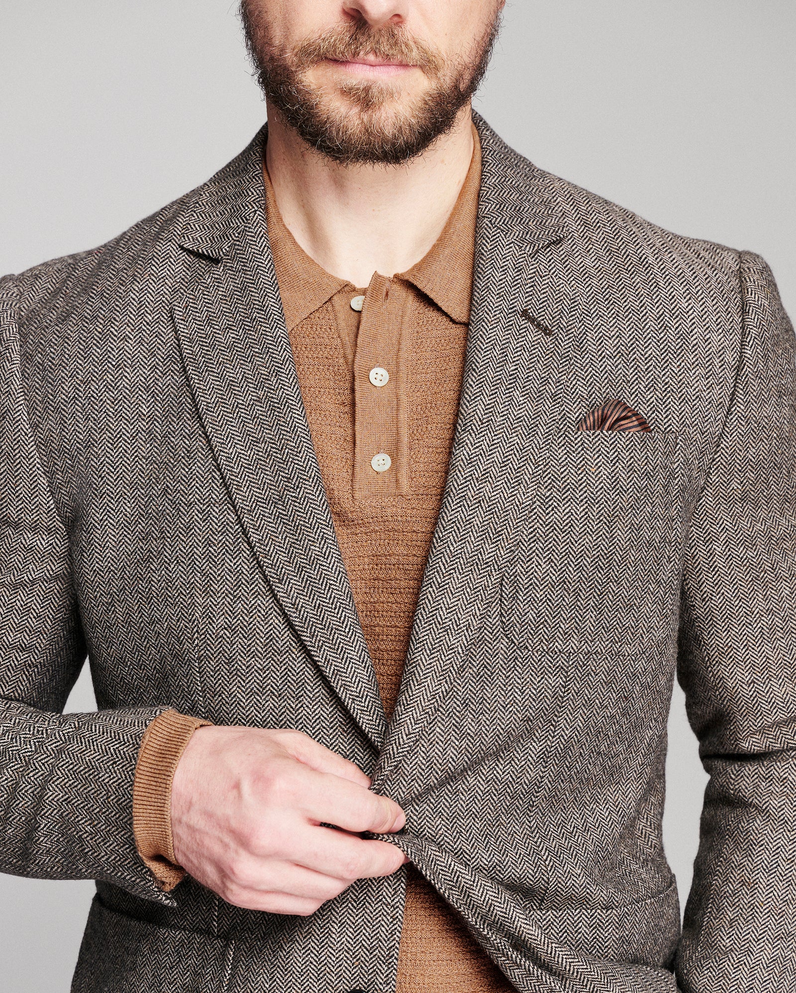 A man wearing a brown and gray herringbone blazer over a brown knit polo shirt, with a matching pocket square, against a solid gray background.