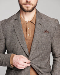 A man wearing a brown and gray herringbone blazer over a brown knit polo shirt, with a matching pocket square, against a solid gray background.