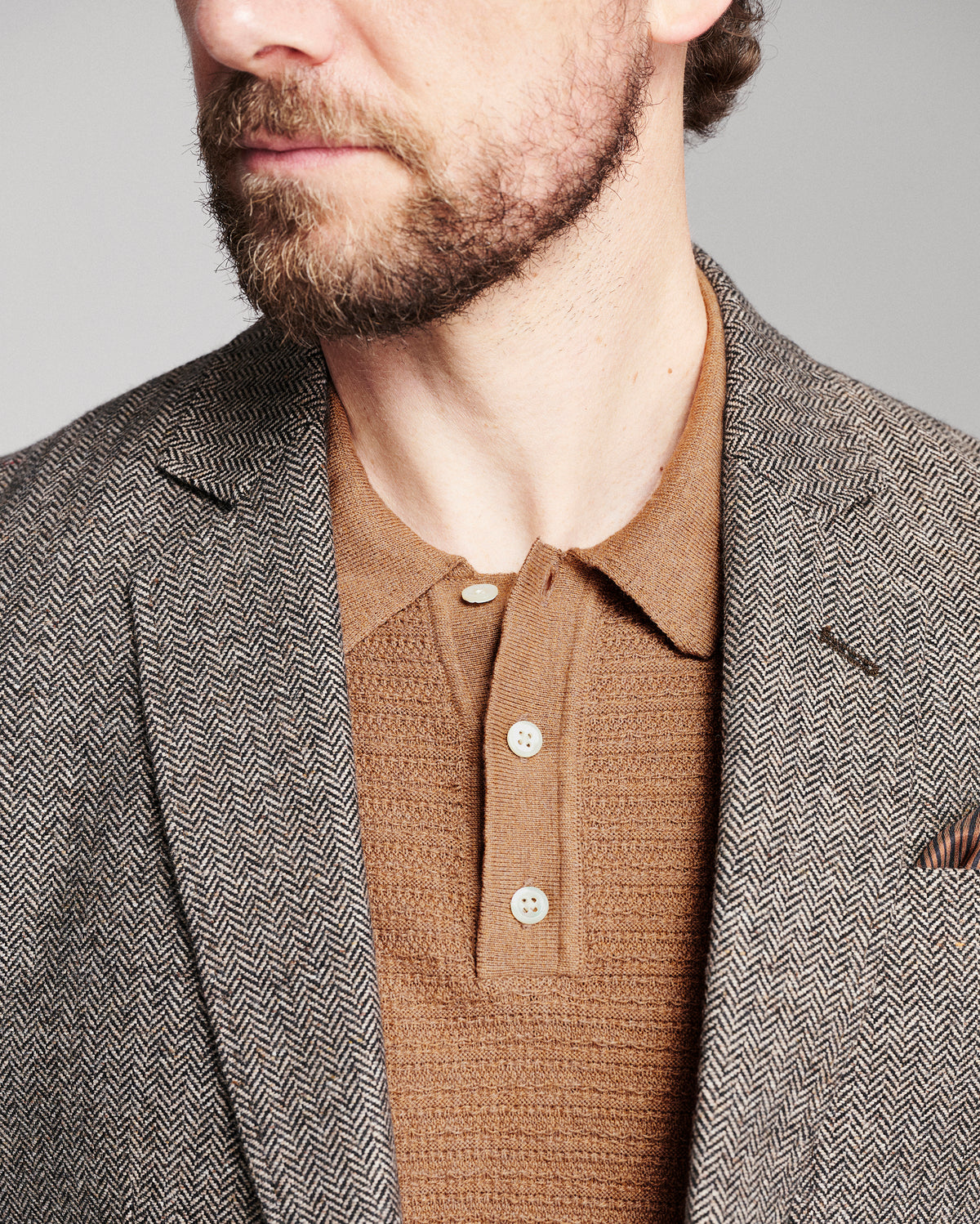 Close-up of a man wearing a brown and gray herringbone patterned blazer over a brown knit polo shirt, against a neutral gray background.