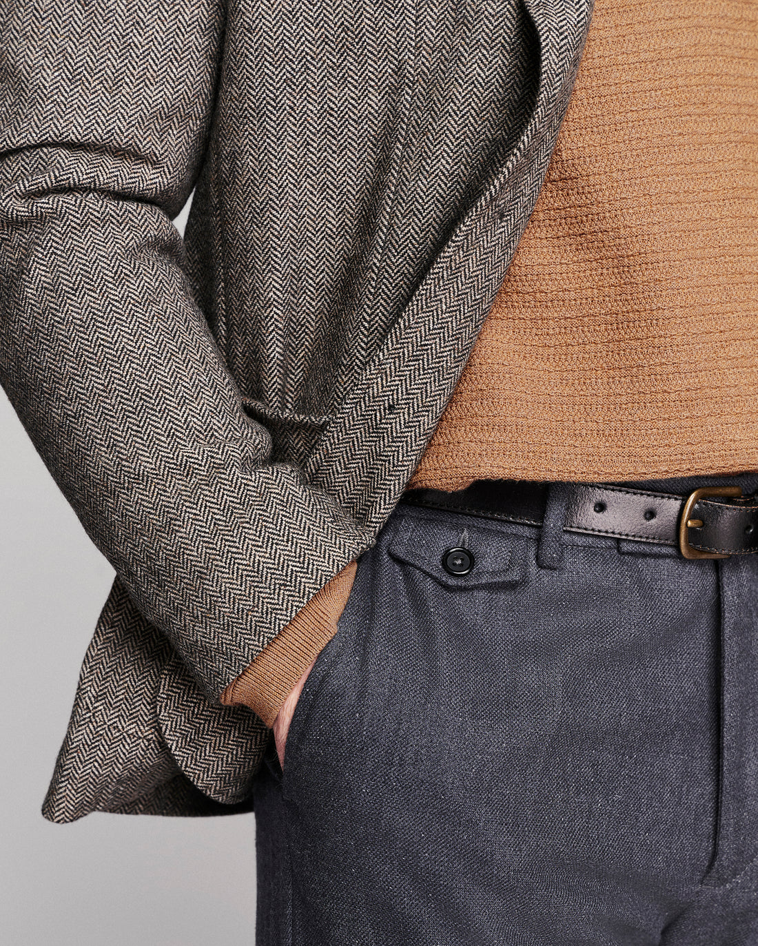 Close-up of a person wearing a gray and brown herringbone patterned blazer over a tan knit sweater, paired with dark gray trousers and a black belt.