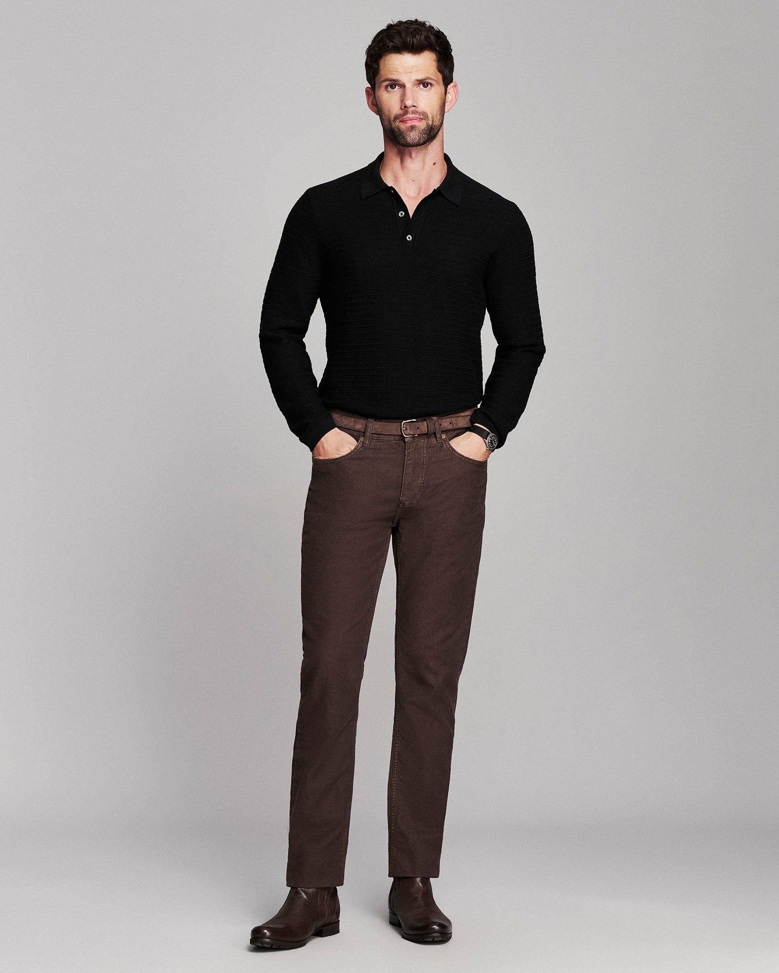 A man modeling a black long-sleeve knit polo shirt and brown trousers against a solid gray studio background.